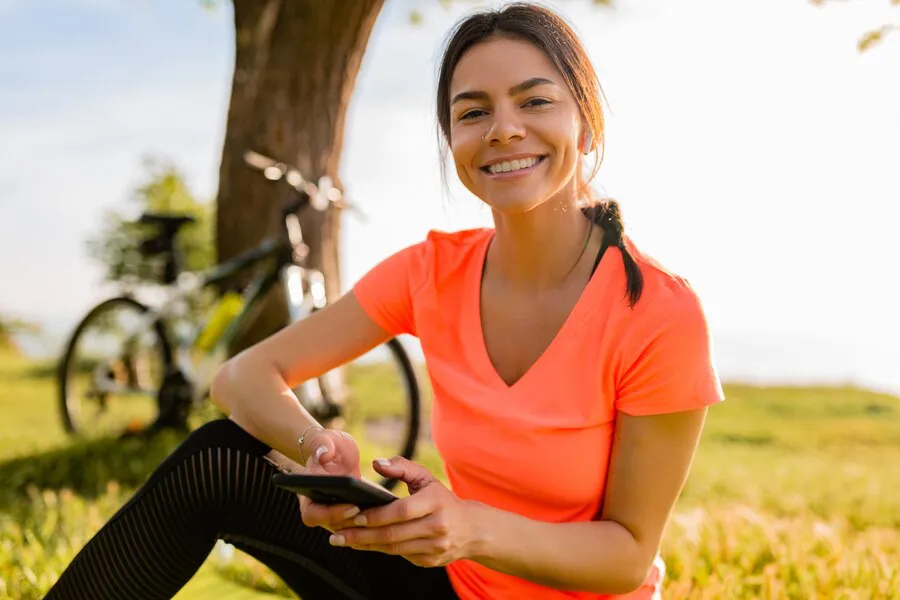 hermosa-mujer-sonriente-sosteniendo-telefono-haciendo-deporte-manana-parque_285396-4290